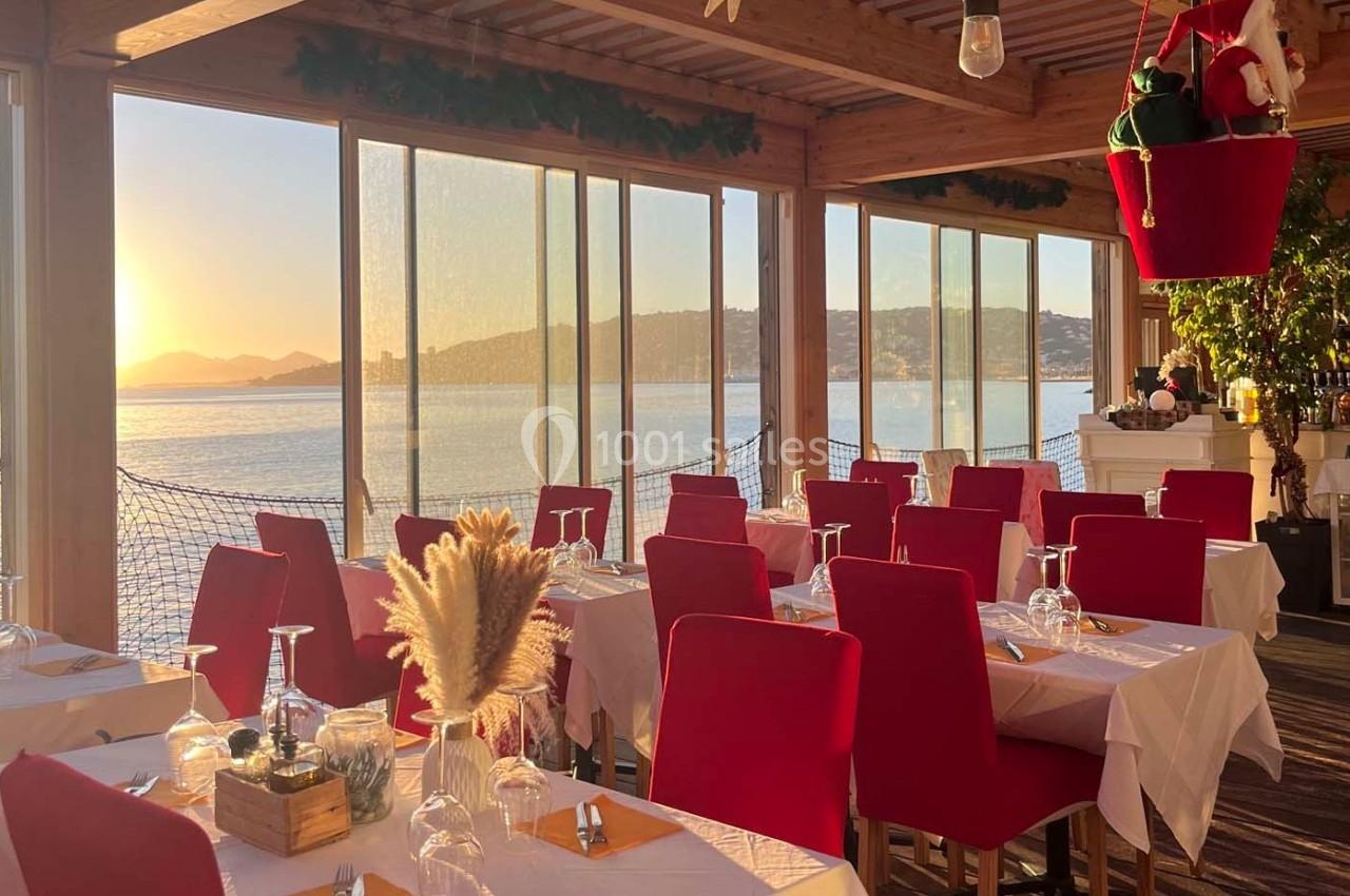 Salle de restaurant lumineuse avec vue sur la mer au coucher du soleil, tables dressées avec nappes blanches et chaises…