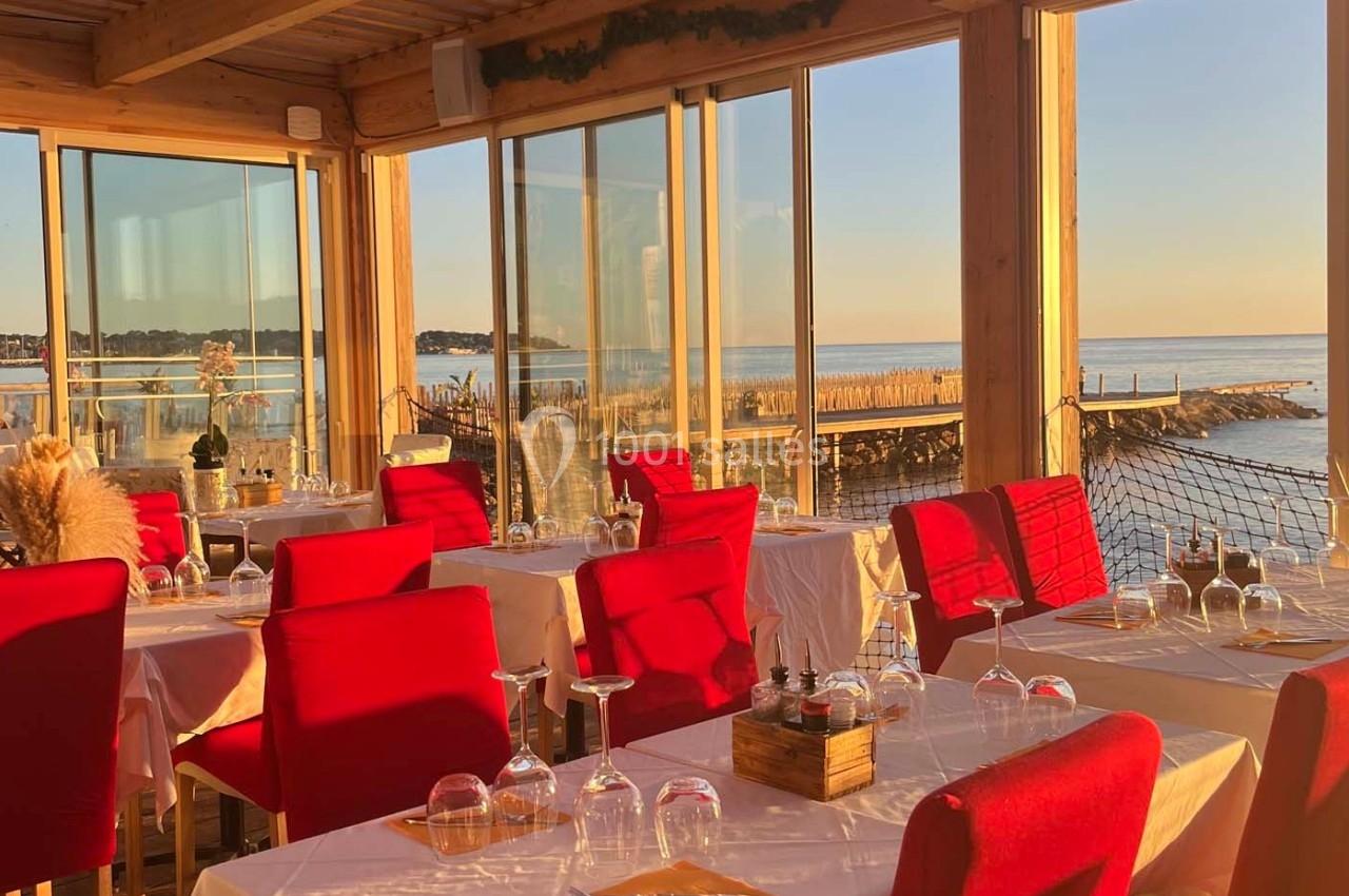 Salle de restaurant lumineuse avec des chaises rouges, vue sur la mer et un ponton au coucher du soleil.