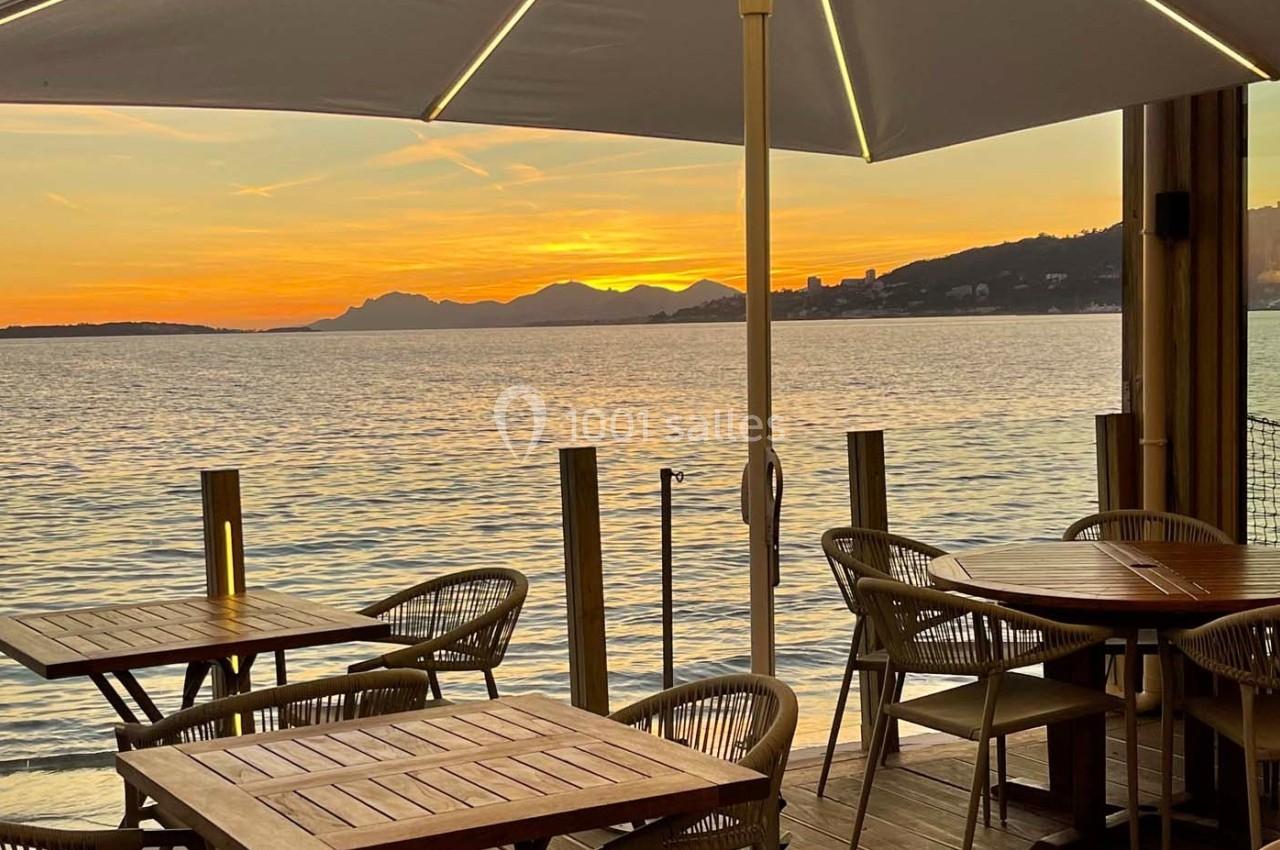 Terrasse en bois avec tables et chaises, vue sur un coucher de soleil au bord de l'eau et montagnes en arrière-plan.