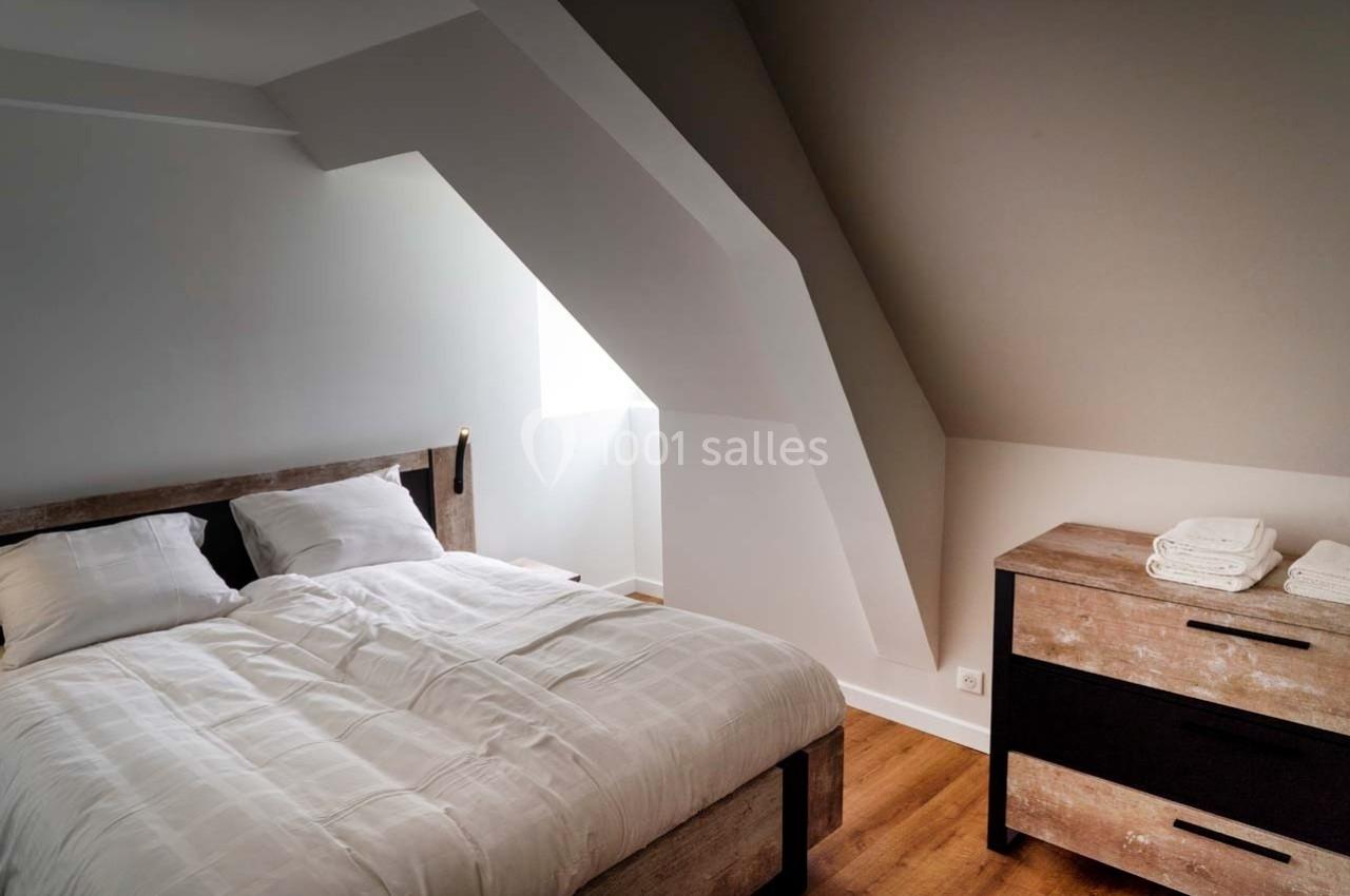 Chambre mansardée avec un lit double, une commode en bois et une fenêtre laissant entrer la lumière naturelle.