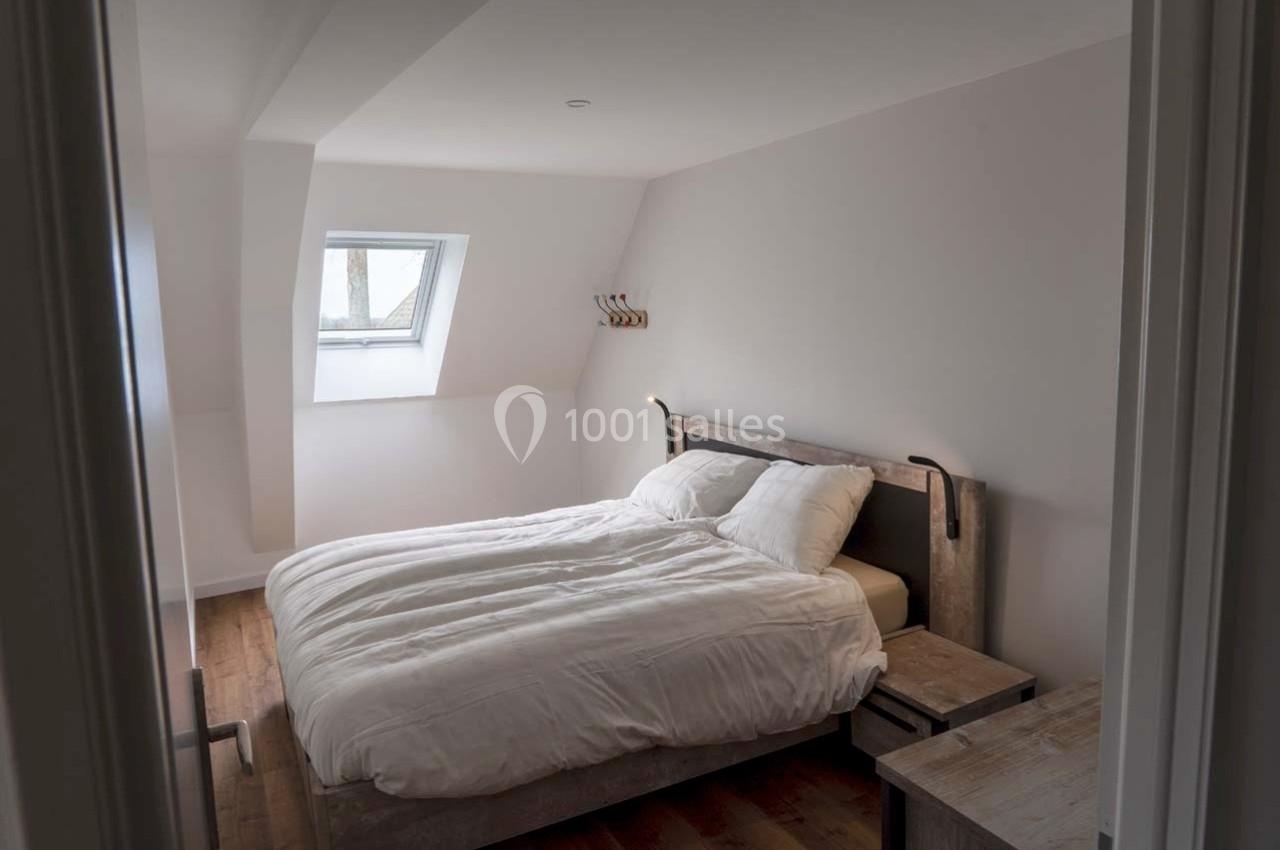 Chambre lumineuse avec lit double, draps blancs, parquet en bois et fenêtre de toit laissant entrer la lumière naturelle.