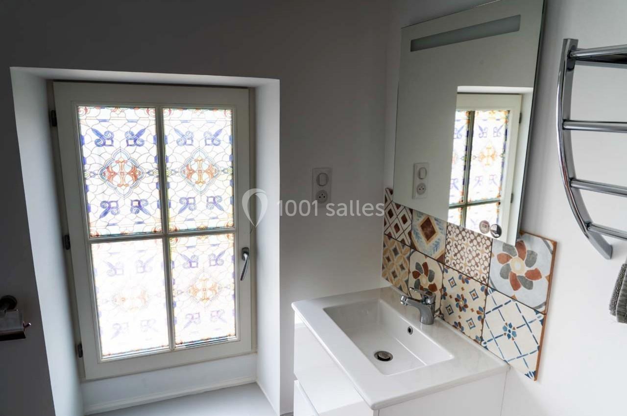 Lavabo blanc avec miroir, carrelage décoratif en mosaïque et fenêtre à motifs colorés dans une salle de bain lumineuse.