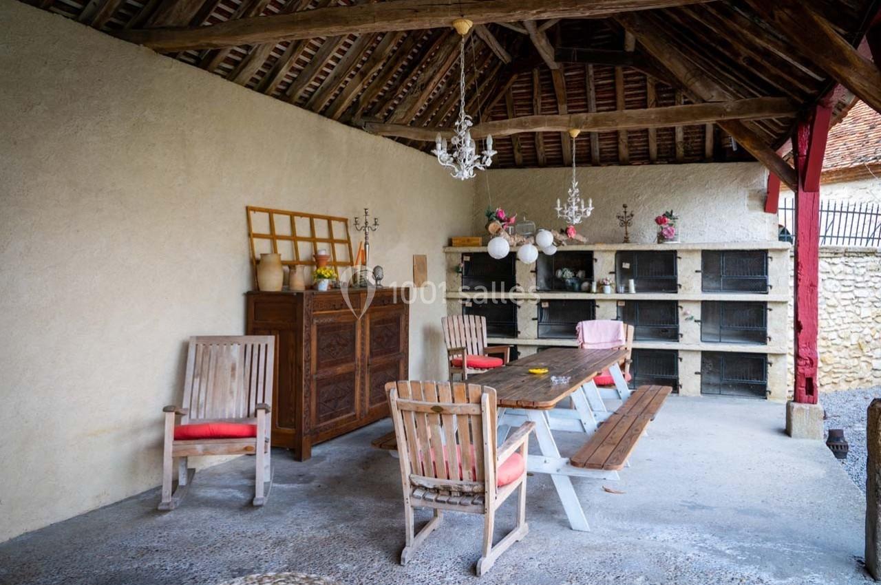 Abri ouvert avec table en bois, chaises, buffet ancien et cages alignées contre un mur sous un toit en poutres apparentes.