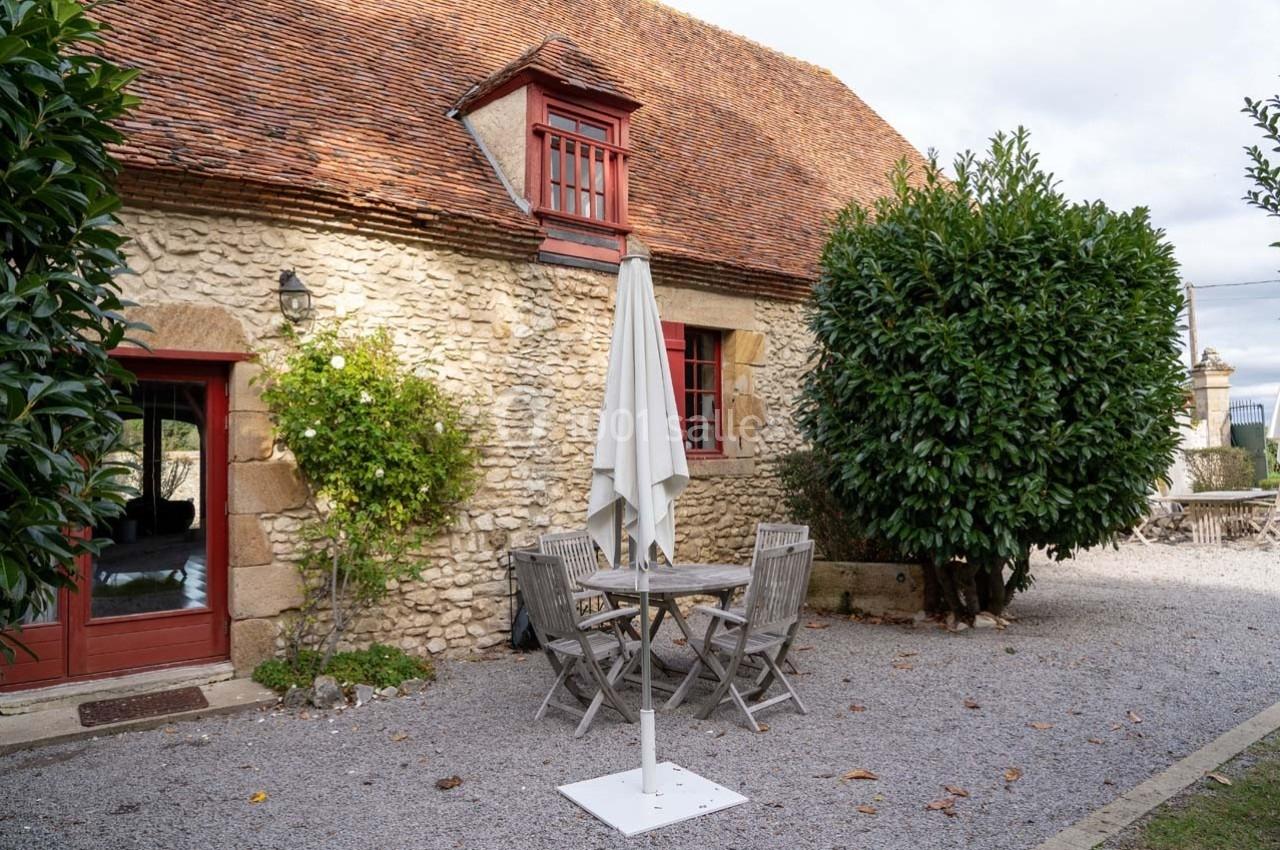 Façade en pierre d'une maison avec toit en tuiles, table et chaises en bois sous un parasol dans une cour gravillonnée.