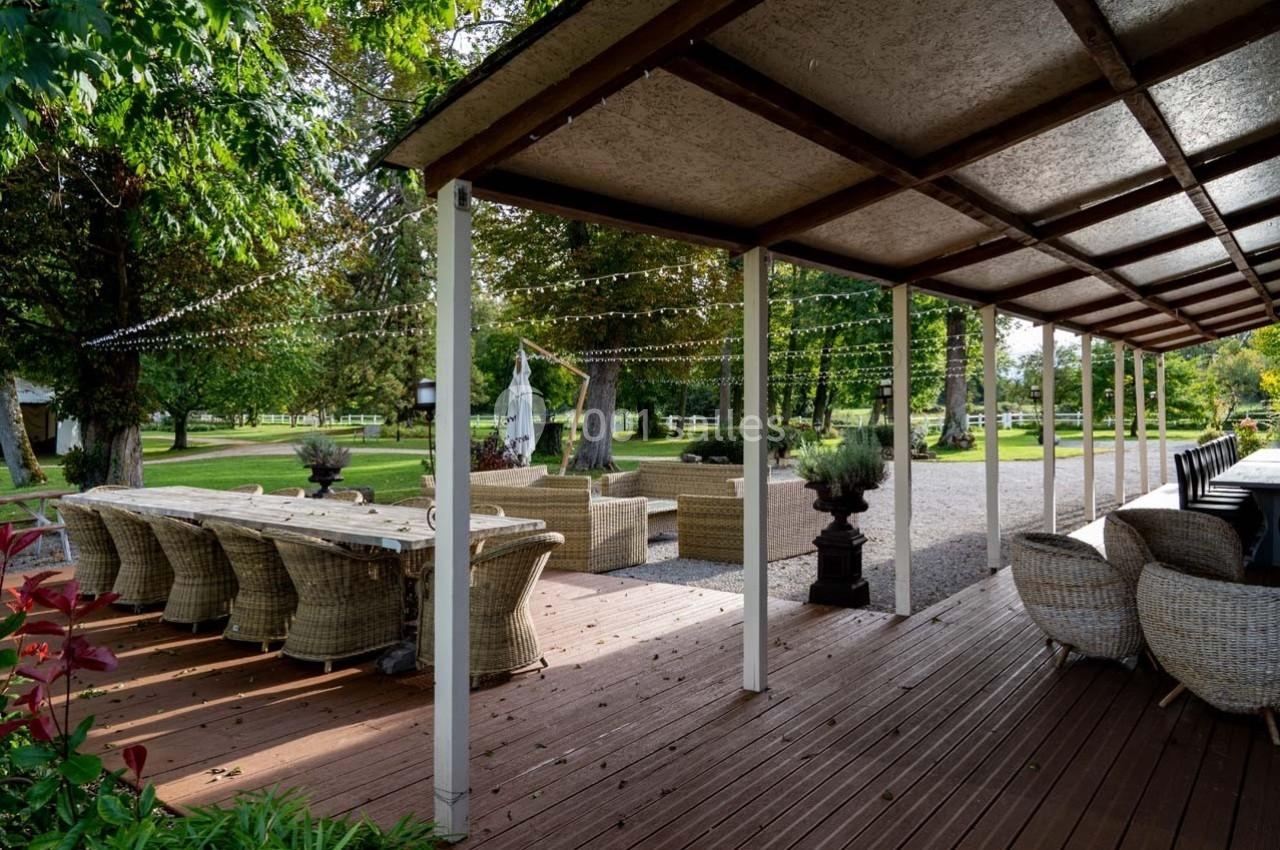 Terrasse en bois avec table et chaises en rotin, surplombée d'un toit, donnant sur un jardin arboré.