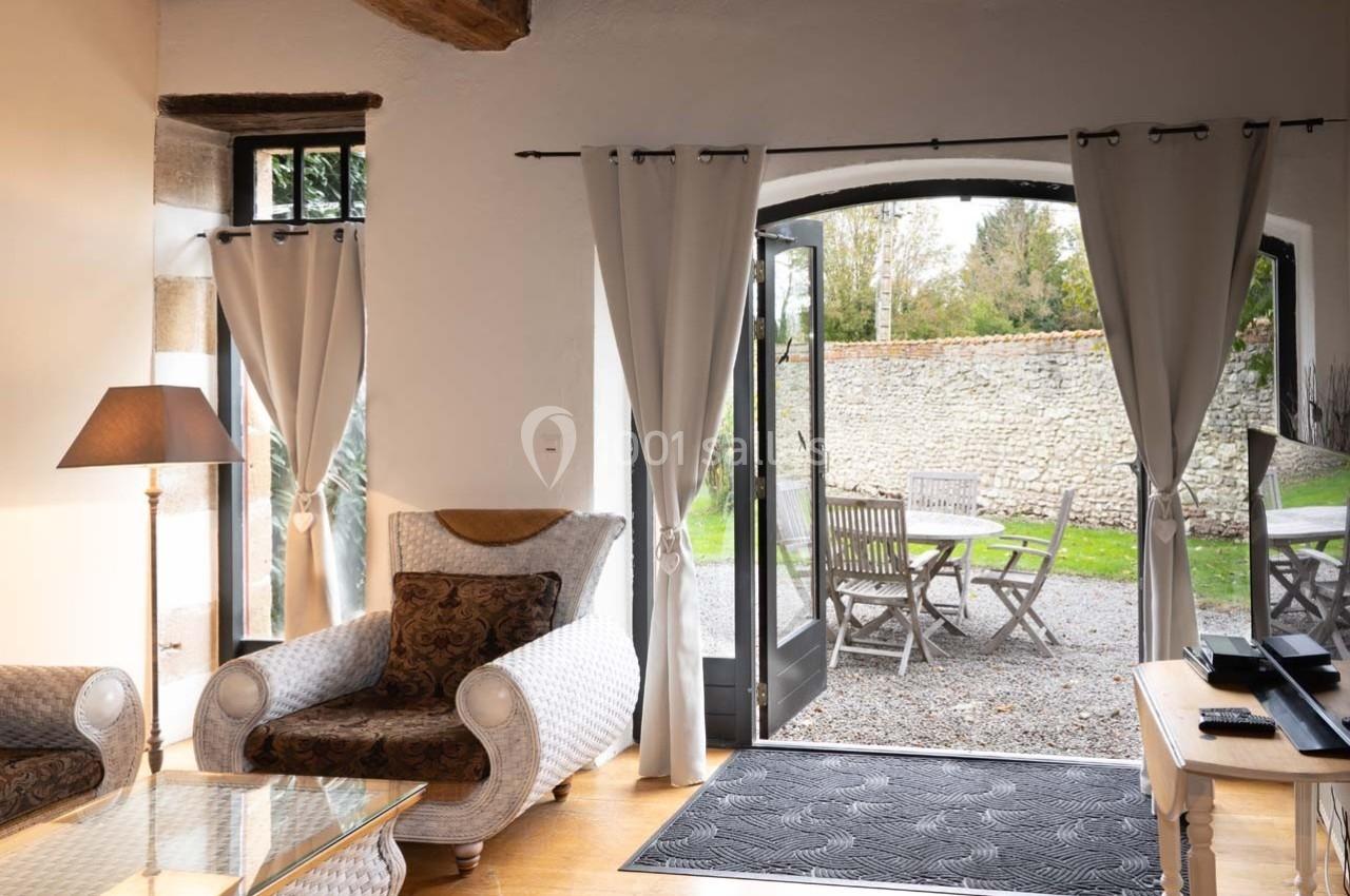 Salon lumineux avec fauteuils, table basse et porte-fenêtre ouverte sur une terrasse avec table et chaises en bois.