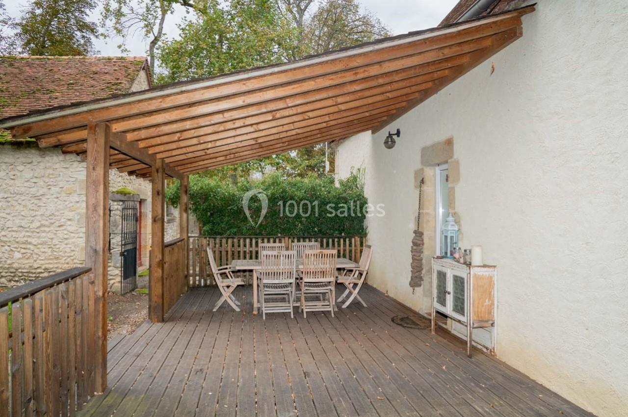 Terrasse en bois couverte avec table et chaises, adossée à une maison blanche entourée de verdure.