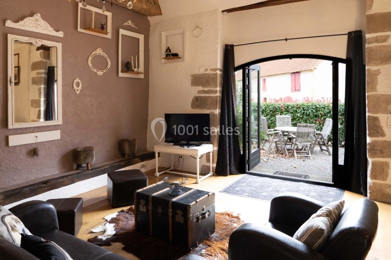 Salon chaleureux avec murs en pierre, mobilier vintage, télévision et vue sur une terrasse extérieure aménagée.