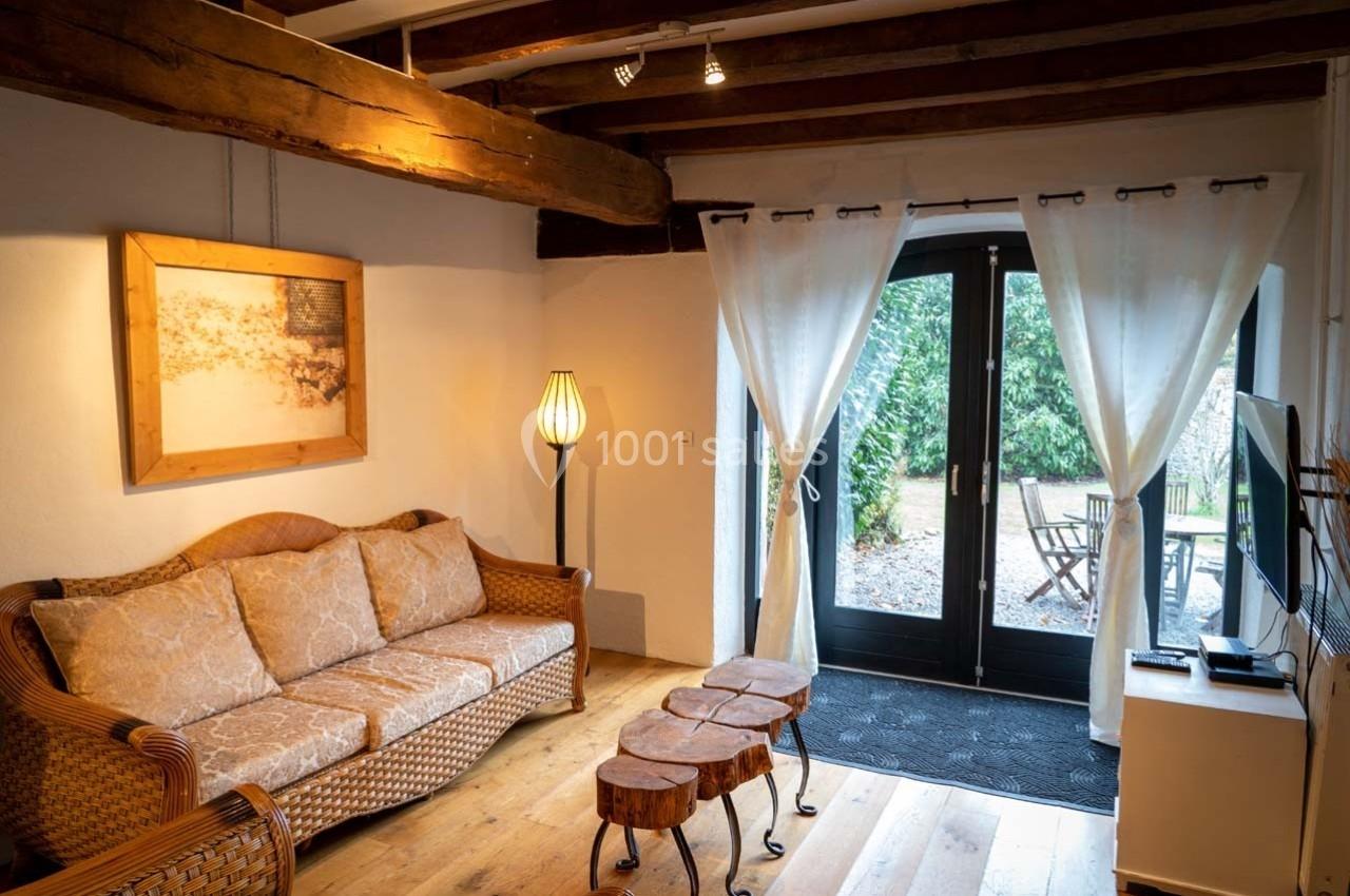 Salon chaleureux avec canapé en rotin, tables en bois, lampadaire, poutres apparentes et vue sur un jardin par une porte…