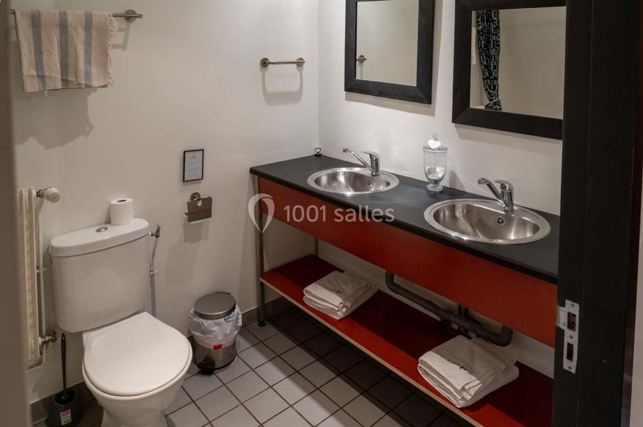 Salle de bain avec toilettes, double vasque, miroirs, serviettes pliées et poubelle sur un sol carrelé.