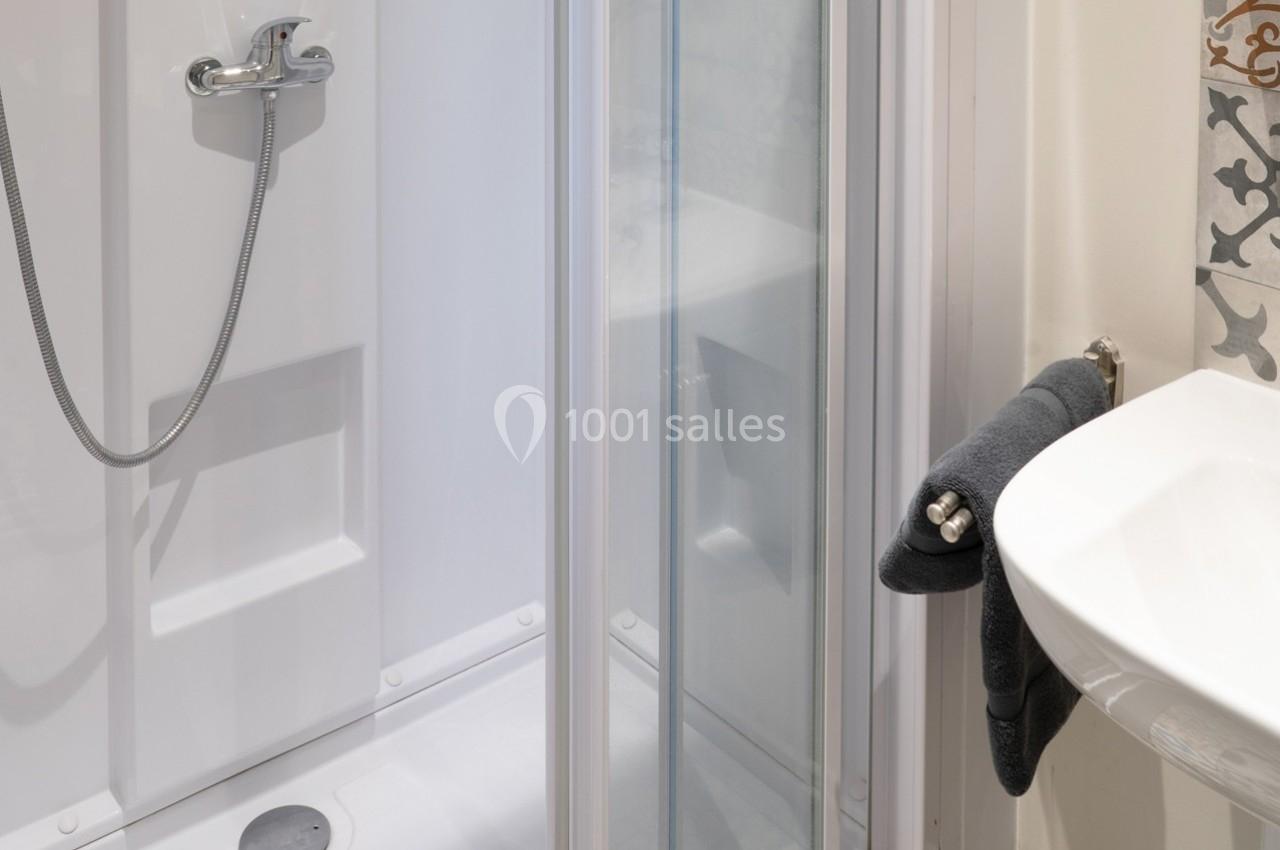 Douche avec paroi vitrée, robinetterie chromée et lavabo adjacent, serviette noire accrochée.