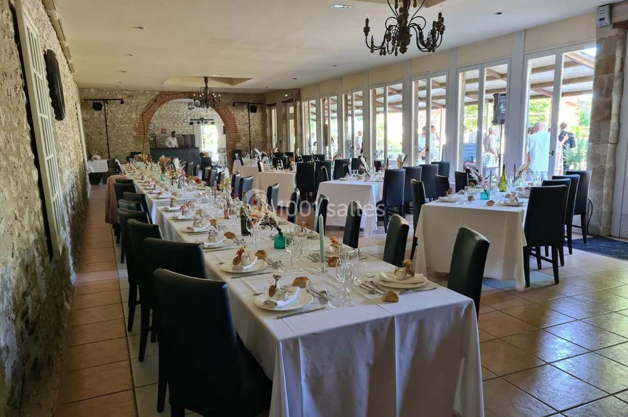 Salle de réception avec tables dressées, nappes blanches, chaises noires et décoration de mariage près de grandes baies…