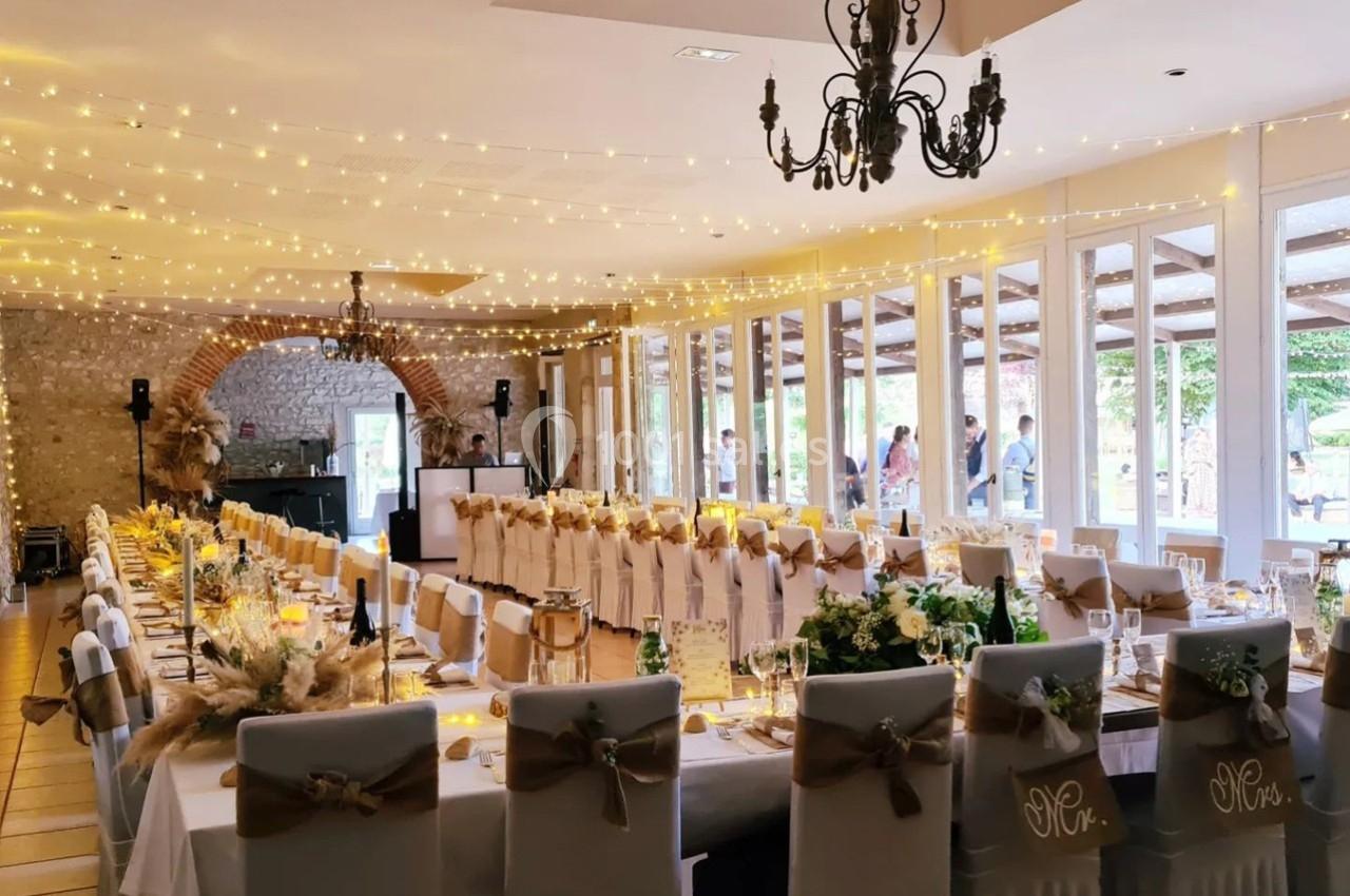 Salle de réception décorée pour un mariage, avec longues tables ornées de nappes blanches, guirlandes lumineuses et chaises…