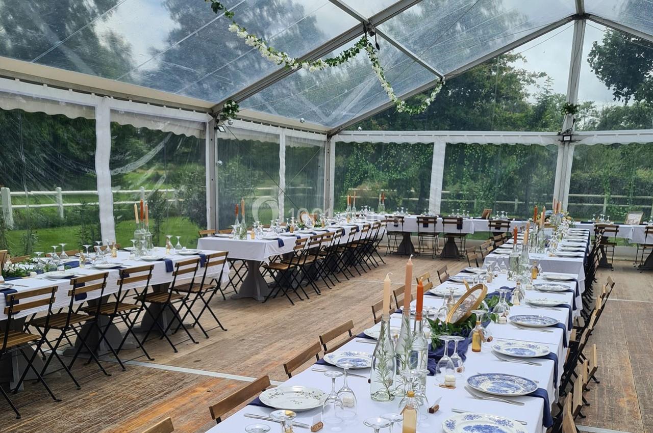 Salle de réception sous une tente transparente, tables dressées avec vaisselle bleue et blanche, décorations végétales.