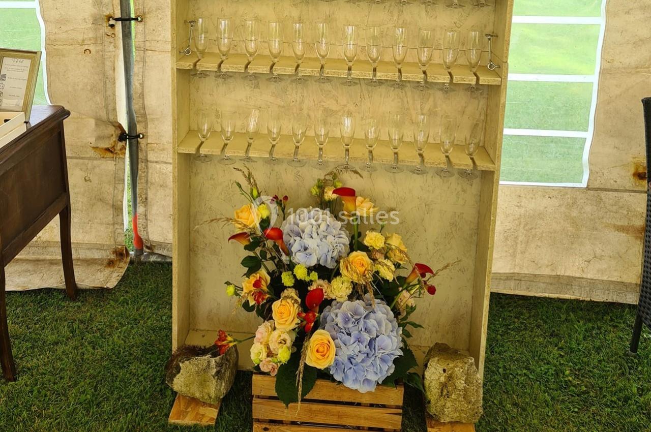 Arrangement floral coloré devant un présentoir en bois contenant des flûtes à champagne, installé sous une tente.