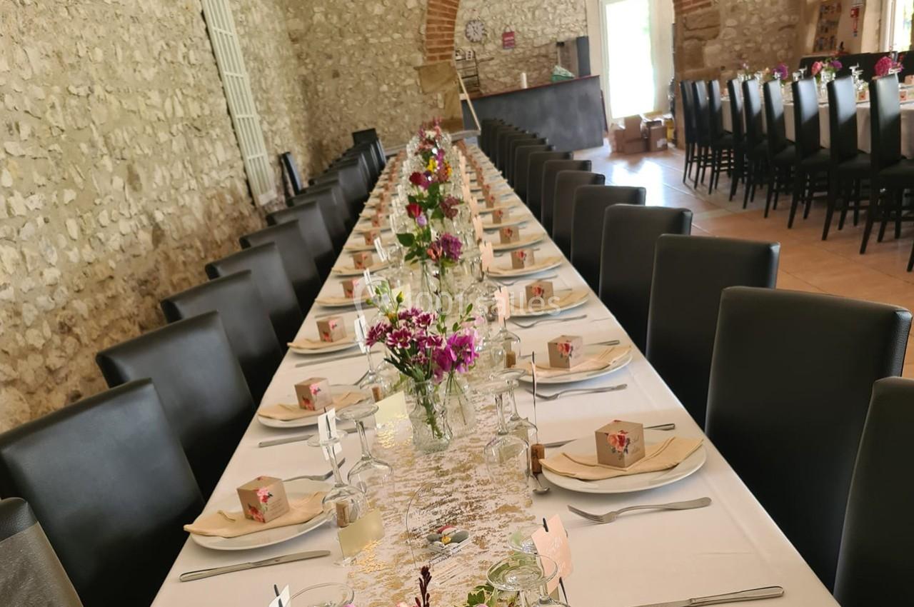 Grande table de banquet décorée de fleurs et de boîtes cadeaux, dans une salle en pierre avec chaises alignées.