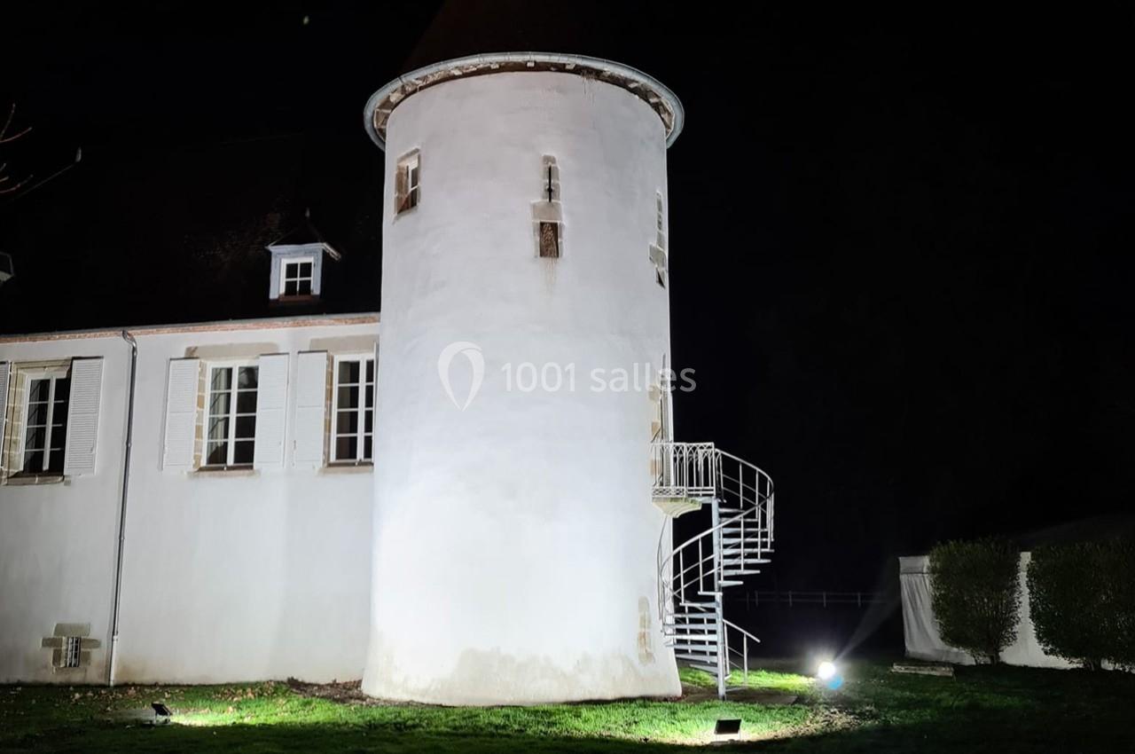 Tour blanche éclairée de nuit avec un escalier en colimaçon extérieur, attenante à un bâtiment aux volets fermés.
