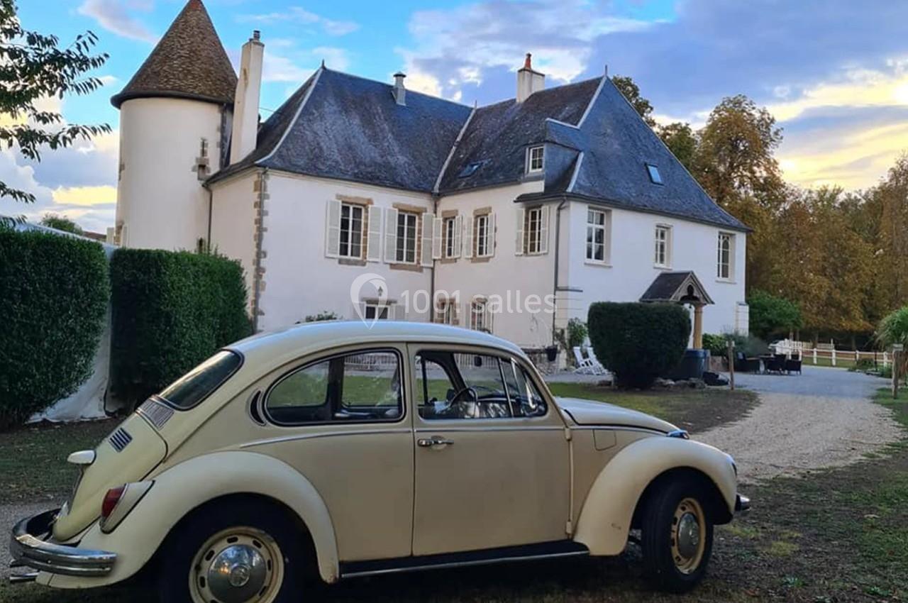 Voiture ancienne beige garée devant une grande maison blanche avec une tour, entourée de verdure.