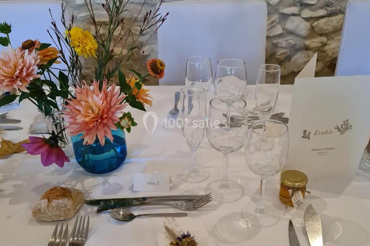 Table dressée avec des fleurs colorées, verres, couverts, menu, pain et un petit pot de miel sur une nappe blanche.