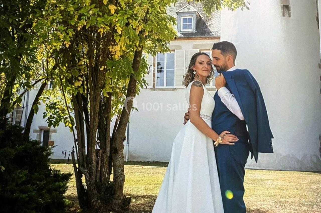 Un couple en tenue de mariage pose sous un arbre devant un bâtiment blanc avec des volets ouverts.