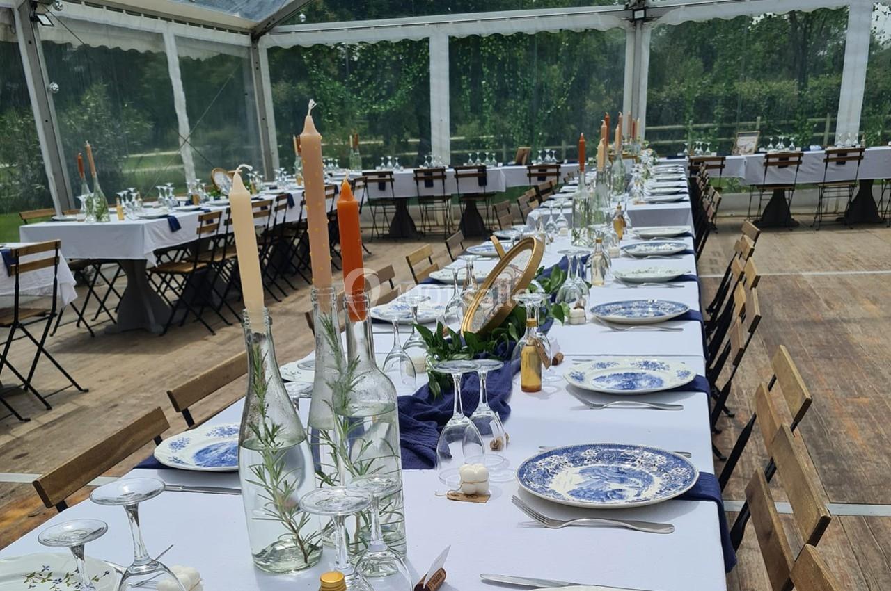 Tables dressées avec nappes blanches, vaisselle en porcelaine bleue et bougies, dans une tente transparente en extérieur.