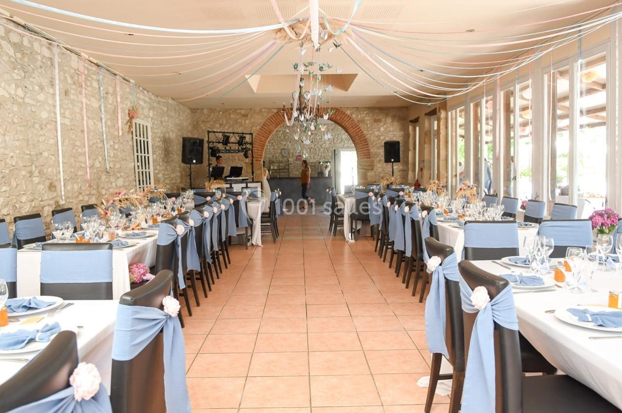Salle de réception décorée avec des tables alignées, nappes bleues, chaises ornées et guirlandes suspendues.