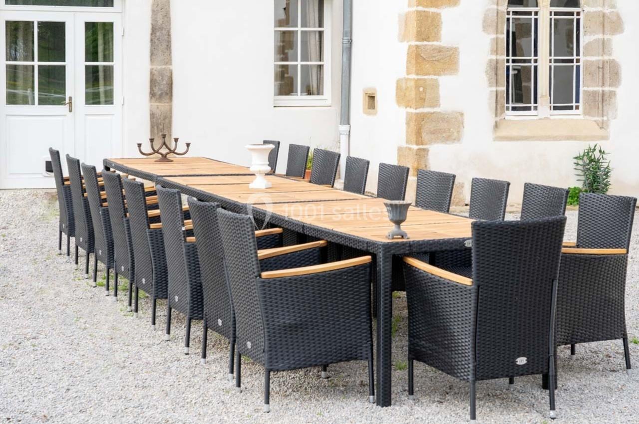Grande table en bois entourée de chaises en rotin noir, installée sur un sol en gravier devant un bâtiment clair.