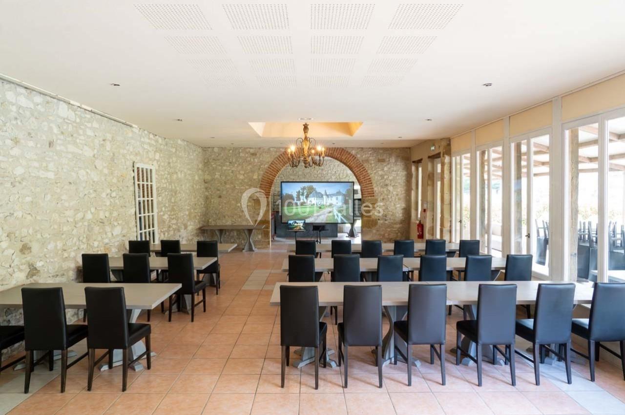 Salle lumineuse avec murs en pierre, tables et chaises noires disposées face à un écran sous une arche en briques.