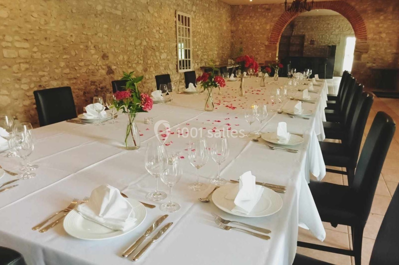 Grande table rectangulaire dressée avec nappes blanches, vaisselle élégante et décorations florales dans une salle en pierre.