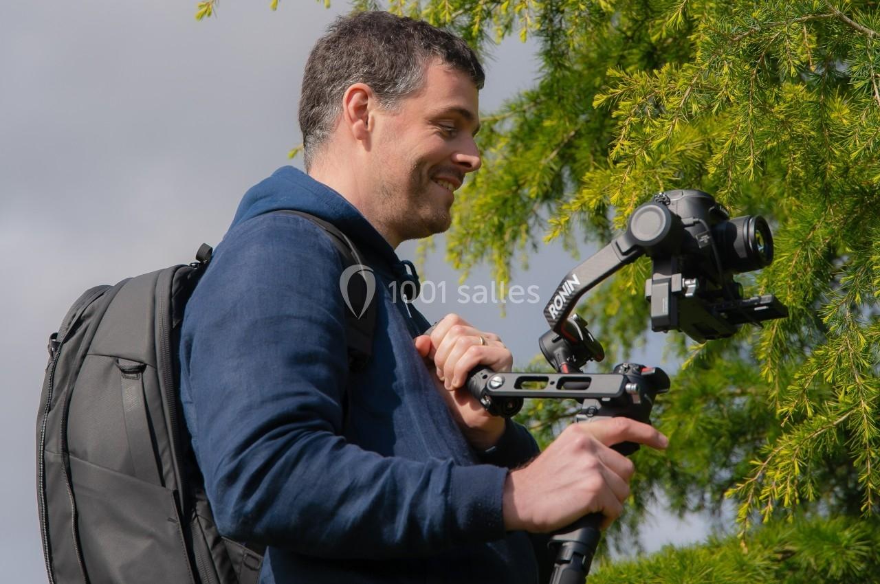 Un homme souriant utilise un stabilisateur avec une caméra, debout à l'extérieur près d'un arbre.
