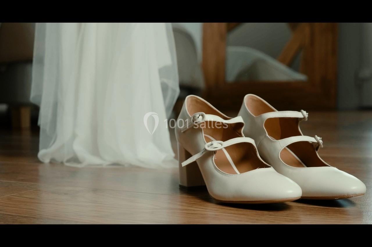 Chaussures blanches à talons posées sur un sol en bois, près d'une robe en tissu léger.