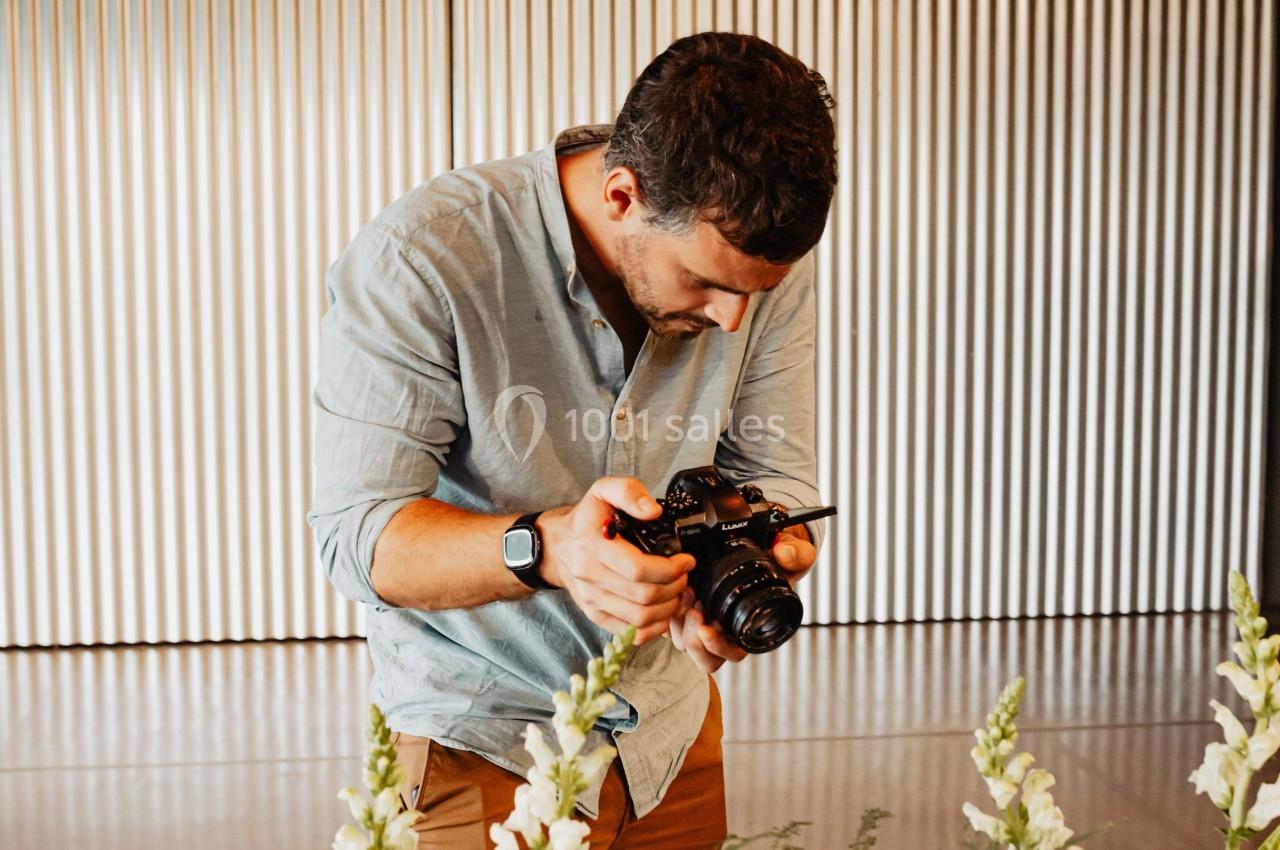 Un homme photographie un arrangement floral sur une table, dans un espace intérieur avec des rideaux métalliques en arrière…