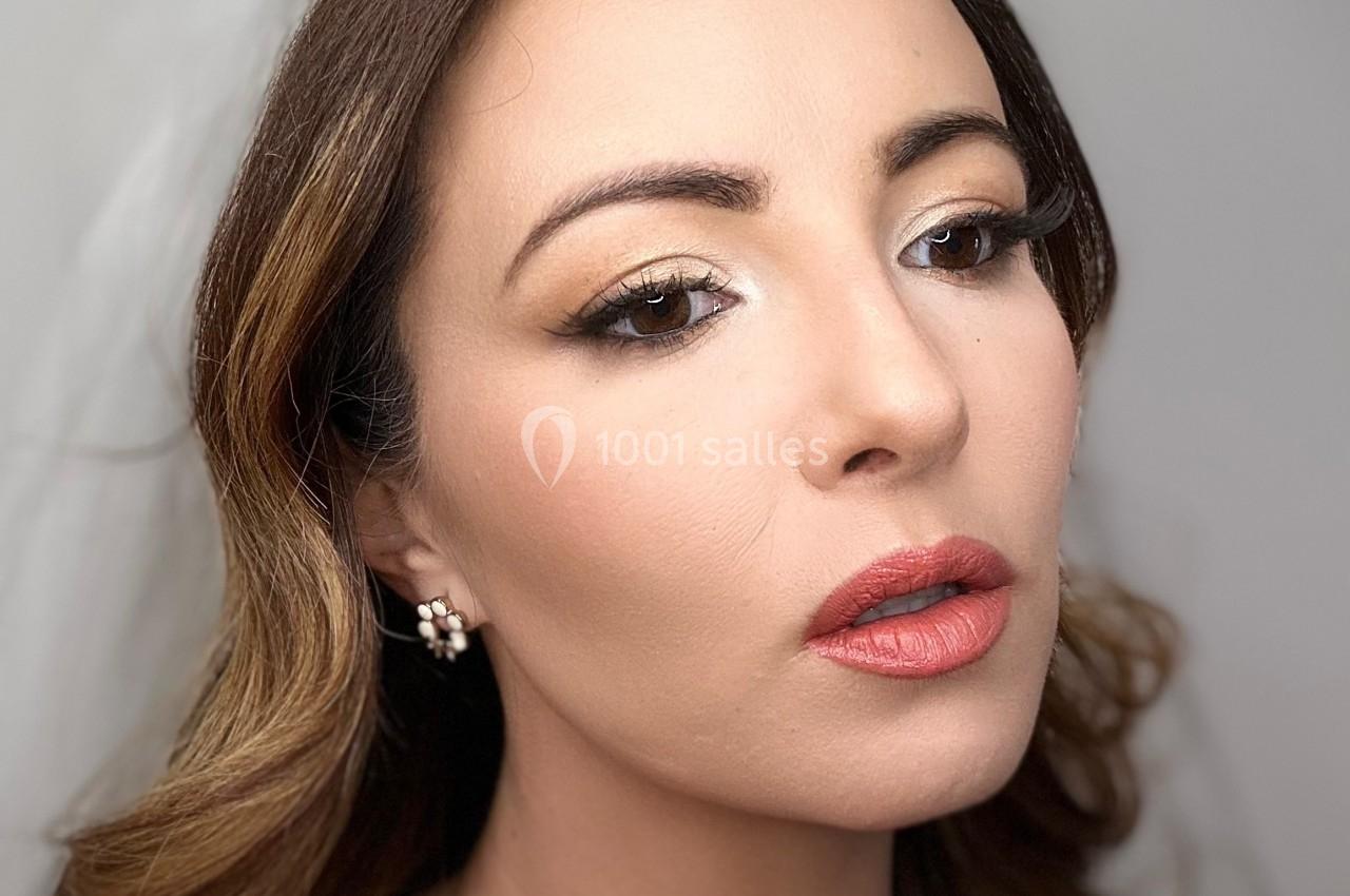 Femme aux cheveux bruns ondulés, maquillage soigné avec rouge à lèvres corail et ombre à paupières claire, fond neutre.