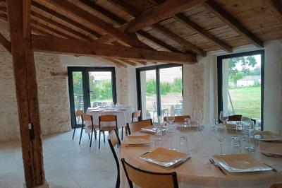 Salle lumineuse avec poutres apparentes, tables dressées et grande baie vitrée donnant sur un extérieur verdoyant.
