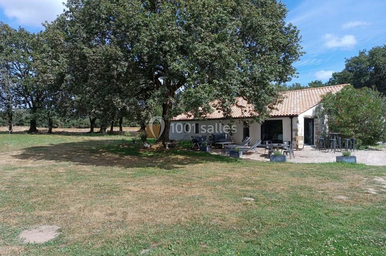 Maison avec toit en tuiles entourée d'arbres, située dans un grand espace herbeux sous un ciel dégagé.