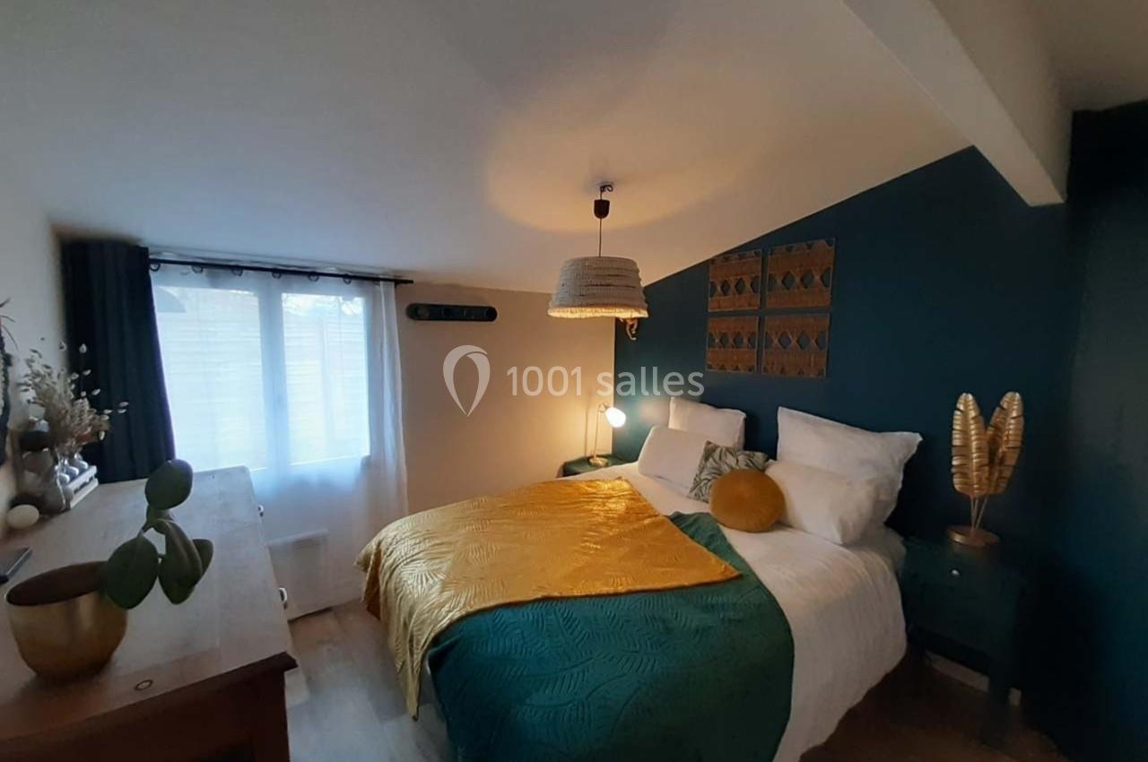 Chambre lumineuse avec lit double, mur bleu, décoration végétale et textile coloré, éclairée par une fenêtre.