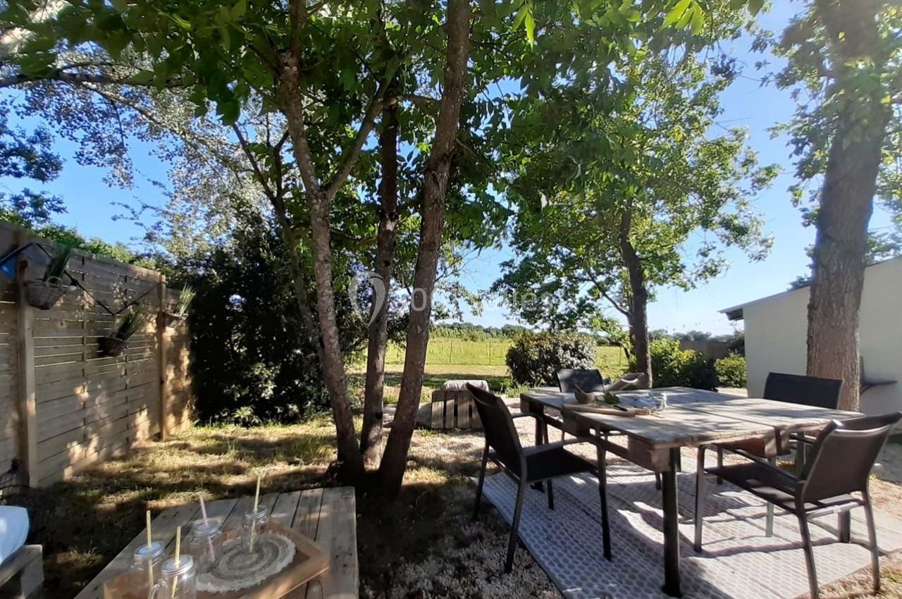 Table de jardin en bois et chaises sous des arbres, avec vue sur un paysage champêtre en arrière-plan.