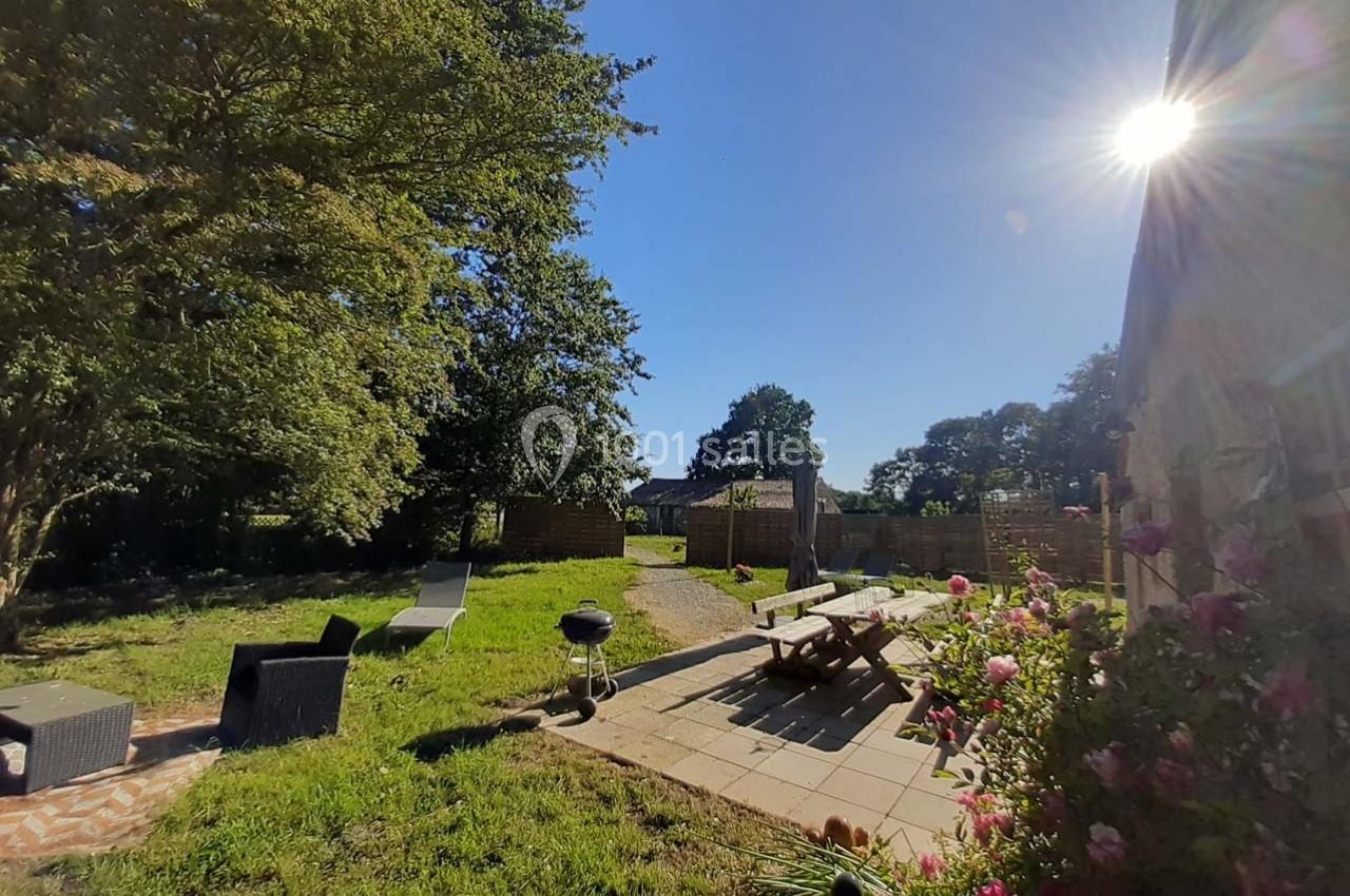 Jardin ensoleillé avec mobilier de jardin, barbecue, table en bois, fleurs et arbres en arrière-plan.