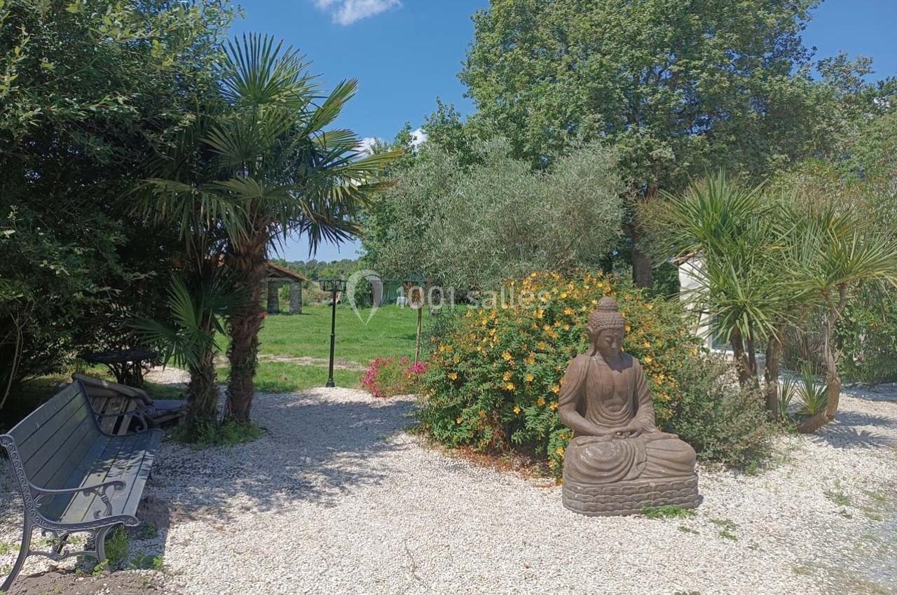 Statue de Bouddha dans un jardin avec banc, palmiers, arbustes fleuris et pelouse en arrière-plan.