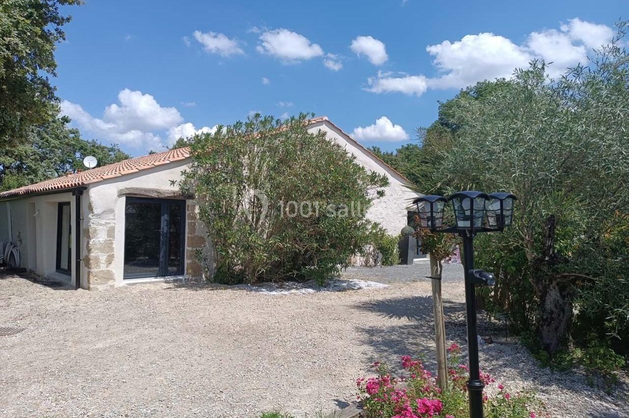 Maison avec façade claire, toit en tuiles, entourée de végétation, allée en gravier et lampadaire au premier plan.