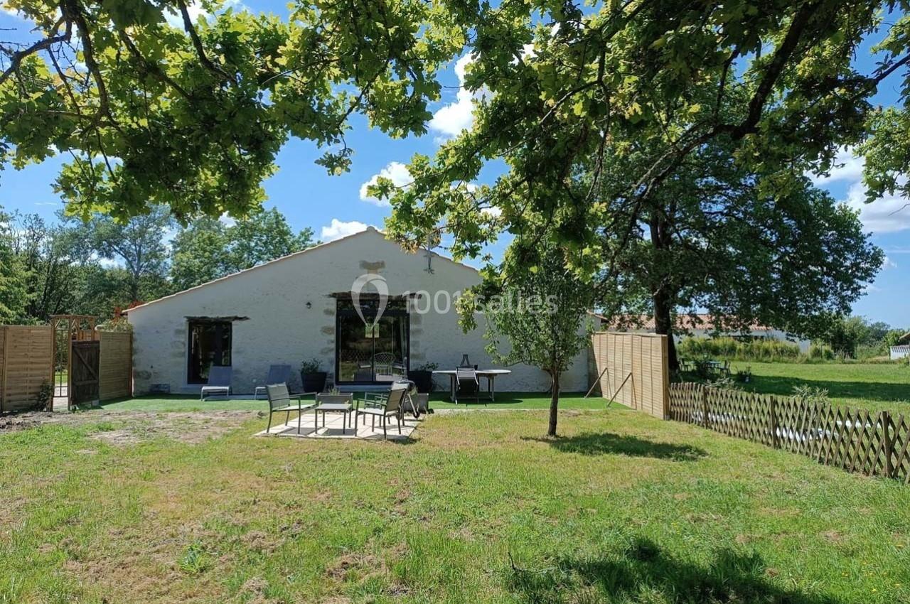 Maison en pierre avec terrasse aménagée, entourée d'un jardin arboré sous un ciel bleu.