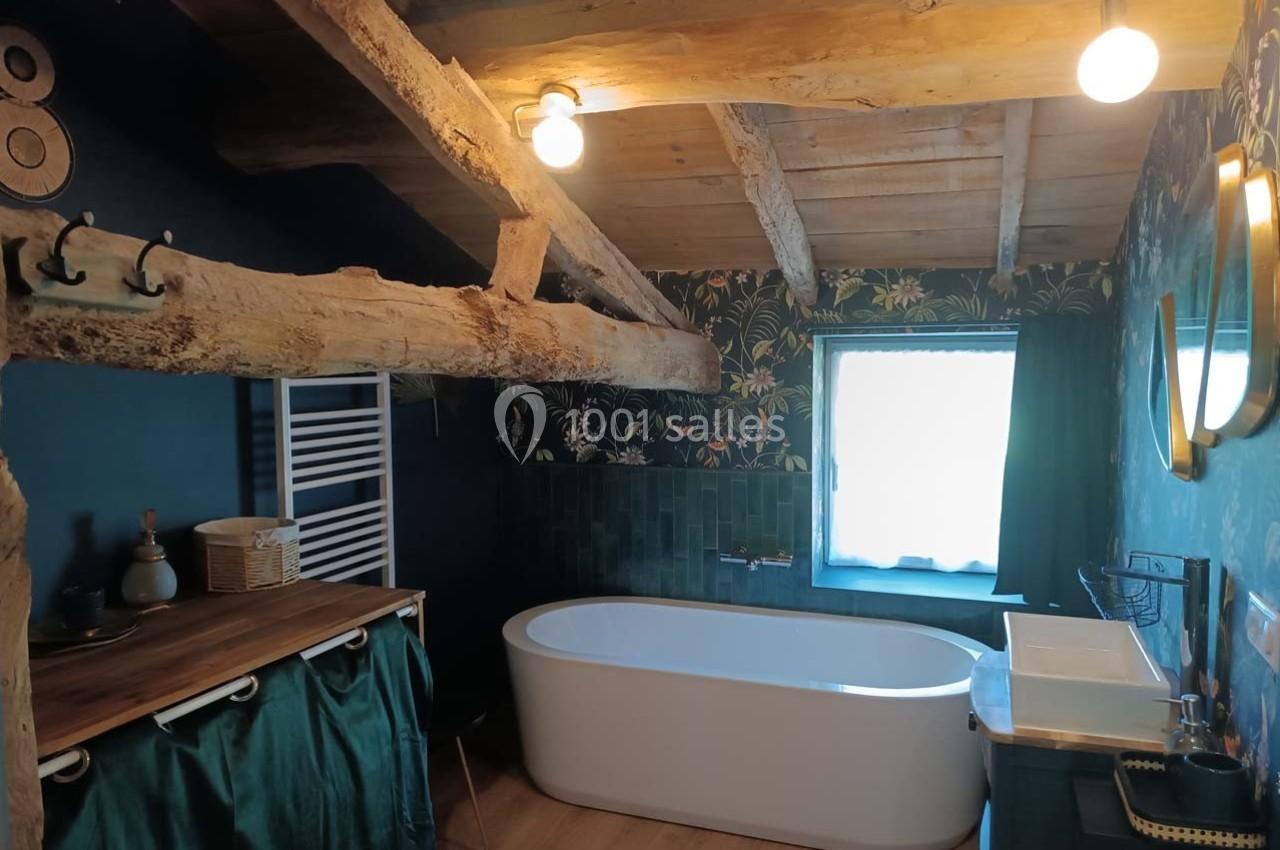 Salle de bain rustique avec poutres apparentes, baignoire blanche, mur bleu foncé et éclairage suspendu.