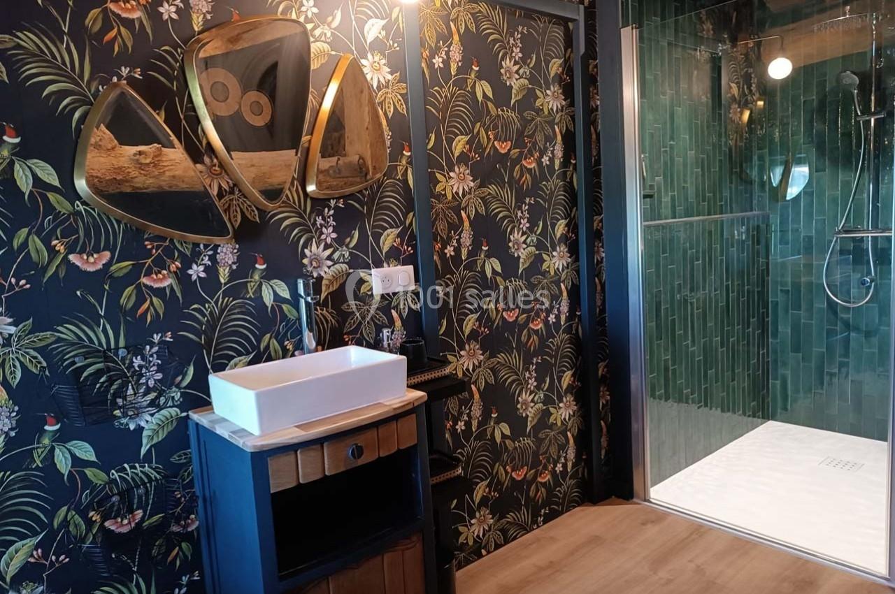 Salle de bain avec lavabo sur meuble en bois, miroir design doré, mur fleuri et douche à paroi vitrée avec carrelage vert.