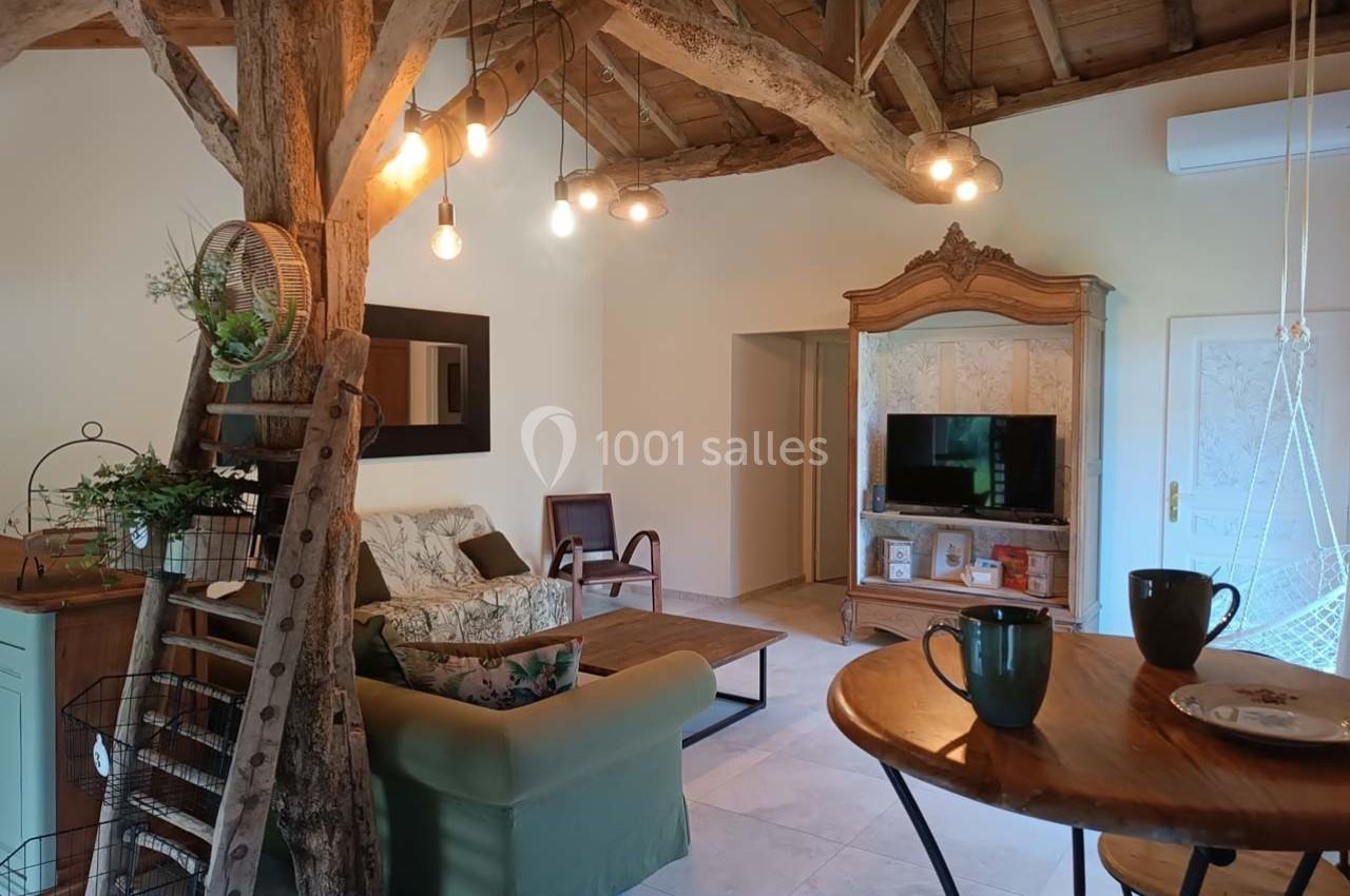 Salon rustique avec poutres apparentes, canapé, meubles en bois, table ronde et éclairage suspendu.