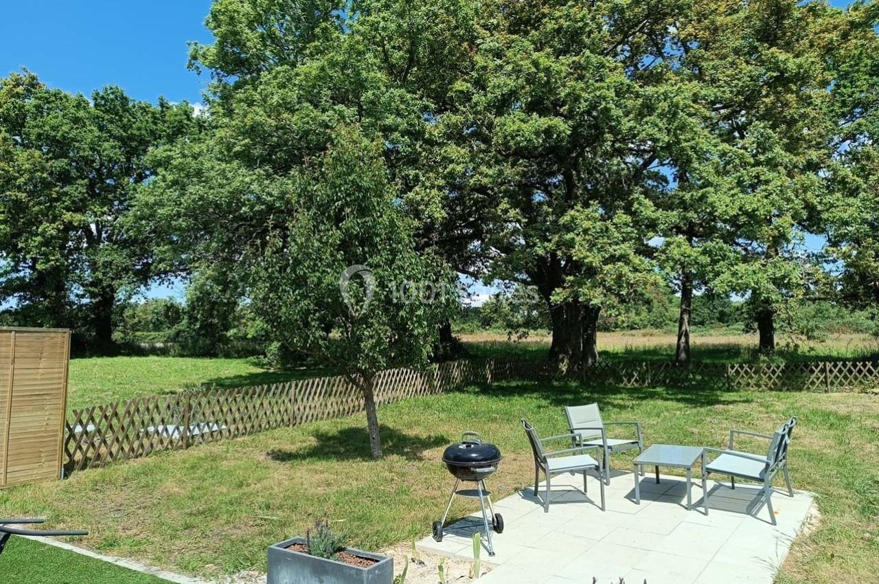 Terrasse avec mobilier de jardin, barbecue et vue sur un jardin arboré entouré d'une clôture en bois.