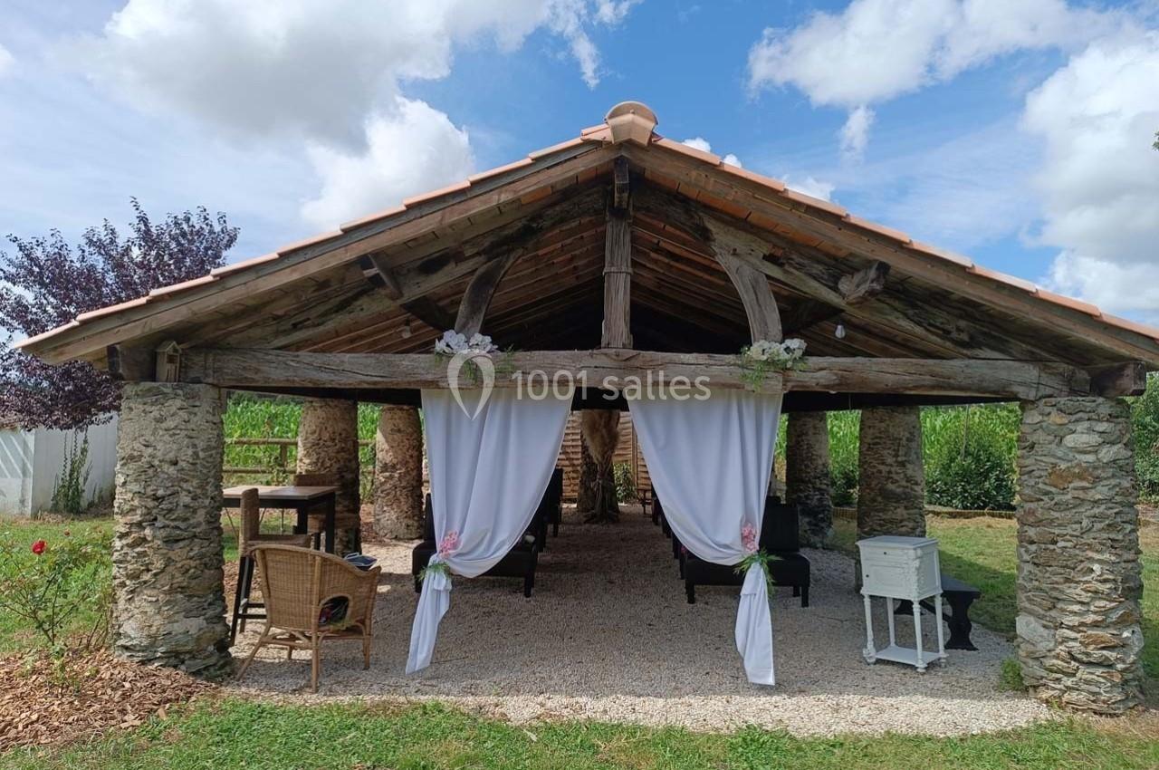 Abri en bois avec toit en tuiles, rideaux blancs décoratifs, mobilier en osier et vue sur un jardin verdoyant.
