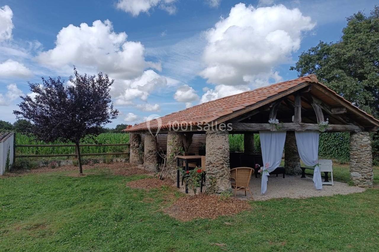 Abri en pierre et bois avec toit en tuiles, rideaux blancs, entouré de verdure et d'un arbre sous un ciel partiellement…