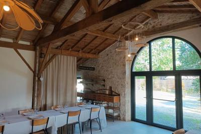 Salle lumineuse avec poutres apparentes, tables dressées et grande baie vitrée donnant sur un extérieur verdoyant.