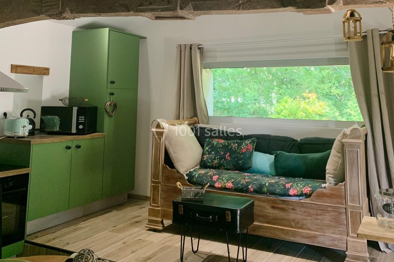 Cuisine ouverte avec meubles verts, canapé en bois avec coussins colorés, grande fenêtre donnant sur un espace arboré.