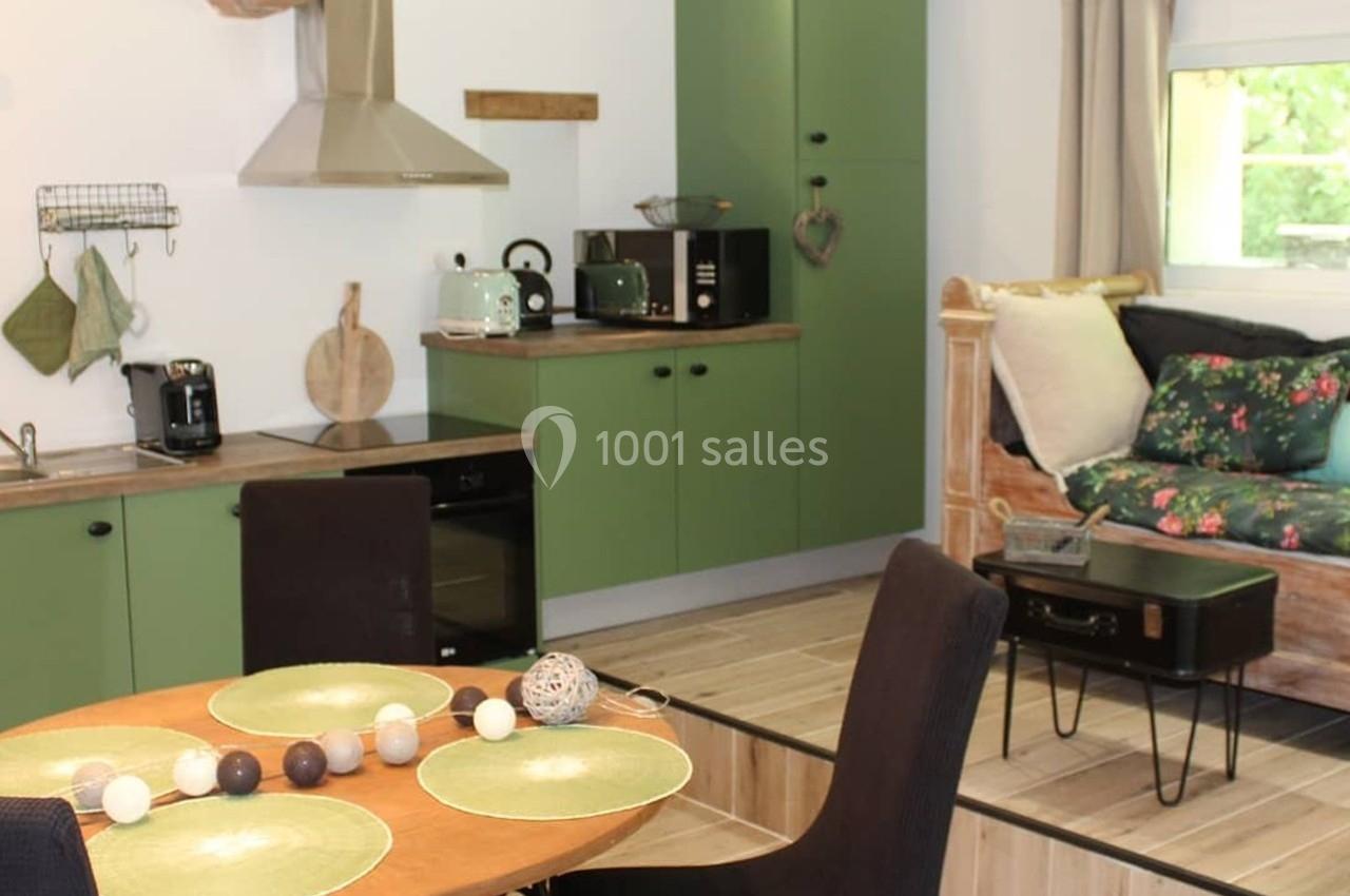 Cuisine ouverte avec meubles verts, table dressée, coin salon avec banquette et fenêtre donnant sur un espace verdoyant.